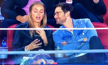 Screenshot of Television captured photo of Liz and Ryan in the stands during the game. Liz is looking at the phone saying something, while Ryan is looking soulfully at her and smiling.